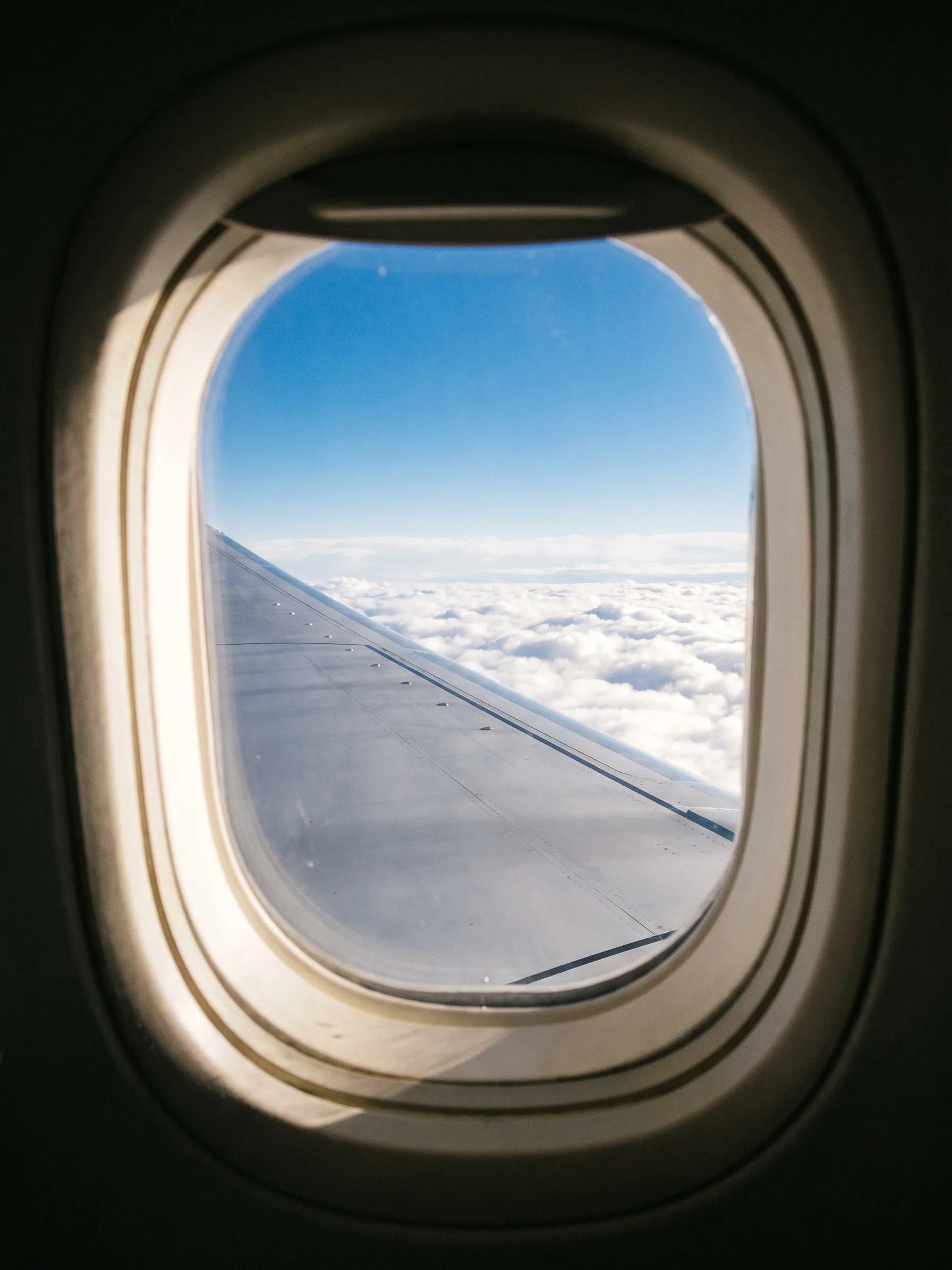 The view through an oval aircraft window: the silver wing over an endless field of white cloud in morning sun